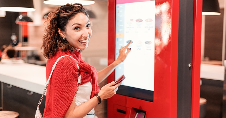 A smiling woman interacts with a self-service ordering kiosk in a fast food restaurant, holding her phone in one hand while selecting items on the touchscreen with the other. She appears cheerful and engaged in the digital ordering process.