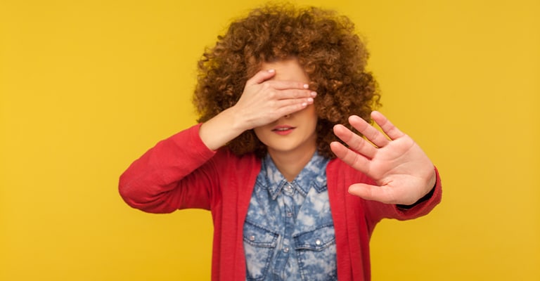 A person with curly hair stands against a bright yellow background, covering their eyes with one hand and holding the other hand out in a gesture that suggests avoidance or reluctance. Their expression and body language convey discomfort or a desire not to see something.