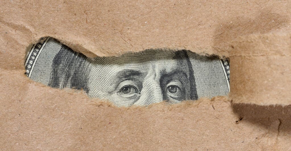 Close up of torn brown paper revealing the eyes of Benjamin Franklin from a US one hundred dollar bill underneath. The ripped opening focuses on the detailed engraved eyes, creating a concept image about hidden money, secrecy, or financial transparency.