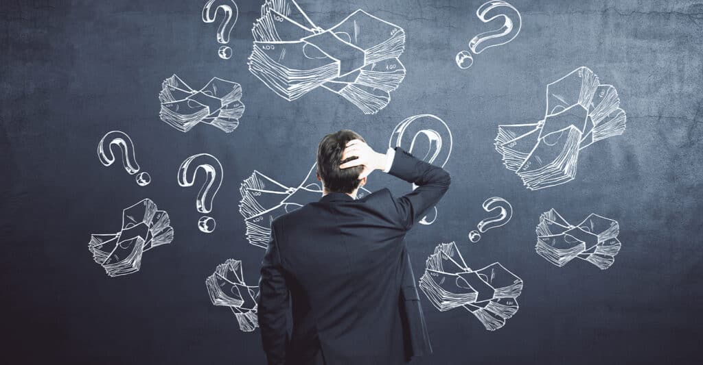 Man in a business suit stands with his back to the camera, scratching his head while facing a chalkboard covered with drawings of cash bundles and question marks. The chalk illustrations of money and uncertainty symbols create a visual concept of financial confusion, budgeting decisions, or money management challenges.