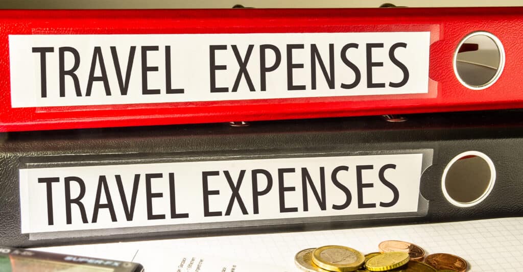 Two office binders labeled TRAVEL EXPENSES sit stacked on a desk, one red and one black, with coins and paperwork partially visible below. The labeled binders clearly organize travel expenses, highlighting record keeping and expense tracking for business or reimbursement purposes.