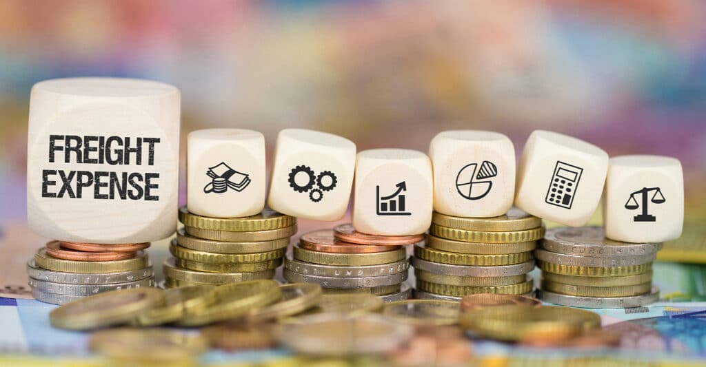 Wooden blocks spelling FREIGHT EXPENSE sit on top of stacked coins, alongside icons of cash, gears, a growth chart, a pie chart, a calculator, and balanced scales. The coins and financial symbols visually represent freight expense costs, budgeting, and financial analysis in logistics and transportation.