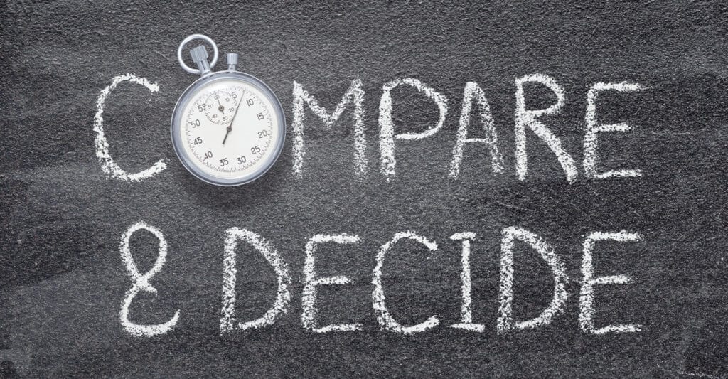 The words COMPARE and DECIDE are written in white chalk on a dark textured surface, with a silver stopwatch placed above the text. The stopwatch and bold lettering visually emphasize the importance of comparing options and making timely decisions.