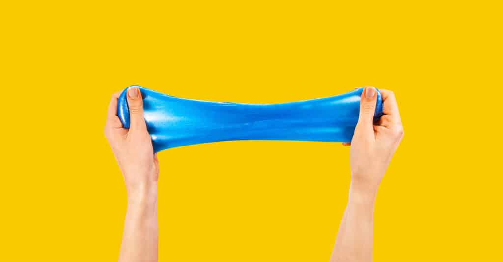 Two hands stretch a piece of bright blue slime horizontally against a solid yellow background. The smooth elastic texture and vibrant colors create a playful visual of sensory play, flexibility, and stress relief.