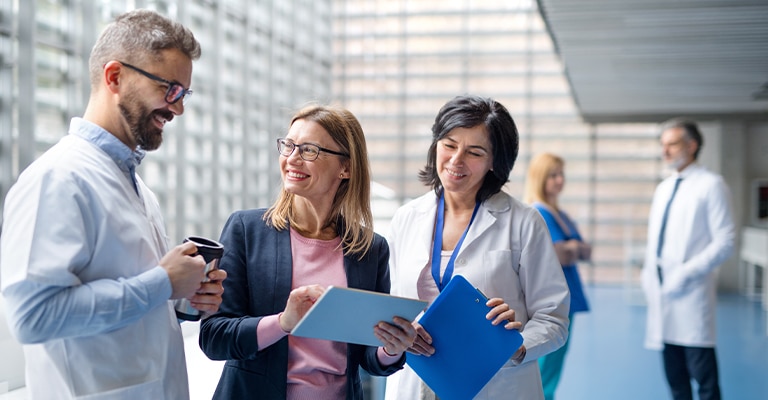 Pharmaceutical Trade Show Hospitality: The Complete Guide 3 Three professionals stand together in a bright hospital or convention corridor, smiling as they review documents and talk. The scene suggests networking or client conversation at a pharmaceutical trade show hospitality setting, with two people in white coats and one attendee in business attire holding folders and a coffee cup.