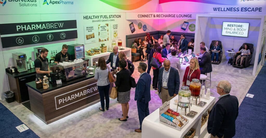a busy trade show booth branded “PHARMA BREW,” featuring a sleek coffee bar with two baristas serving drinks, a healthy snack station with granola bars and infused water, and a lounge area labeled “Connect & Recharge.” Attendees in business attire are mingling, lining up for refreshments, and using massage chairs in a section marked “Wellness Escape” under a sign that reads “Restore: Mind & Body Balanced.